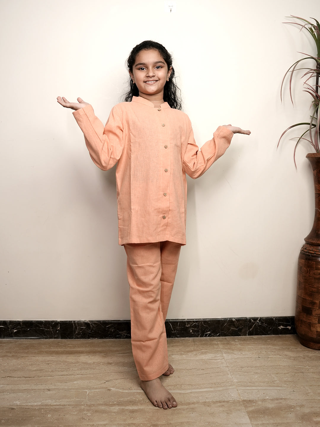 Young girl wearing a peach traditional outfit standing against a plain wall.