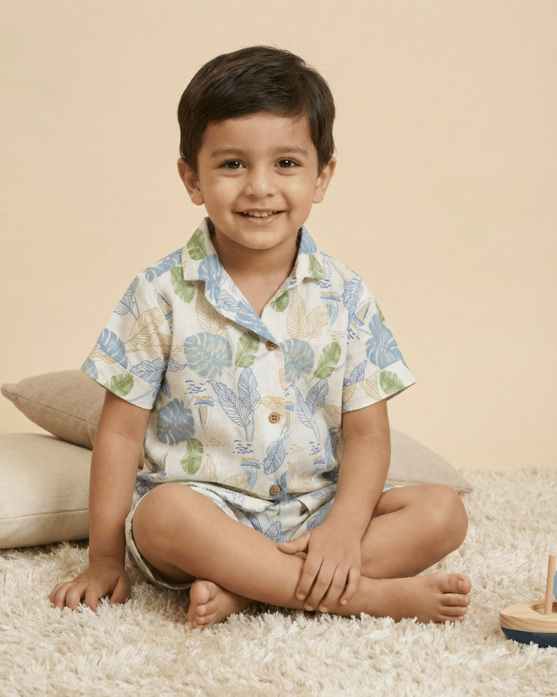 Smiling young boy sitting on a soft rug wearing a tropical leaf-print short pajama set in a cozy room.