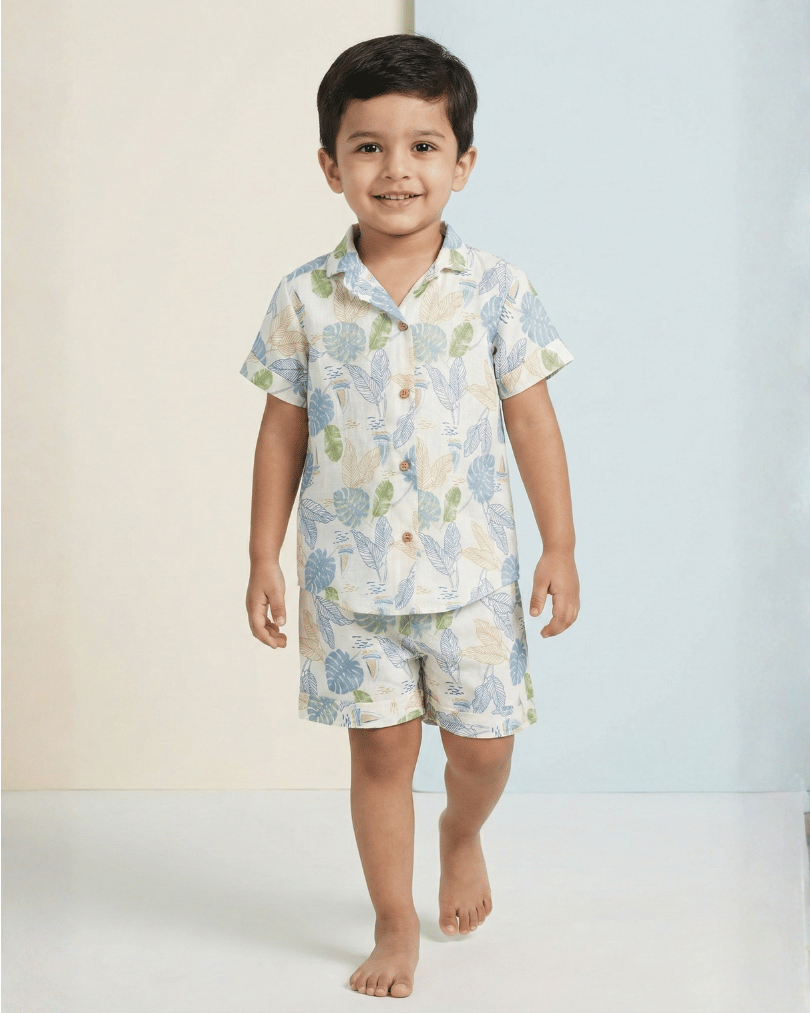 Smiling young boy wearing a lightweight leaf-print pajama set with short sleeves and shorts, walking barefoot on a pastel background.